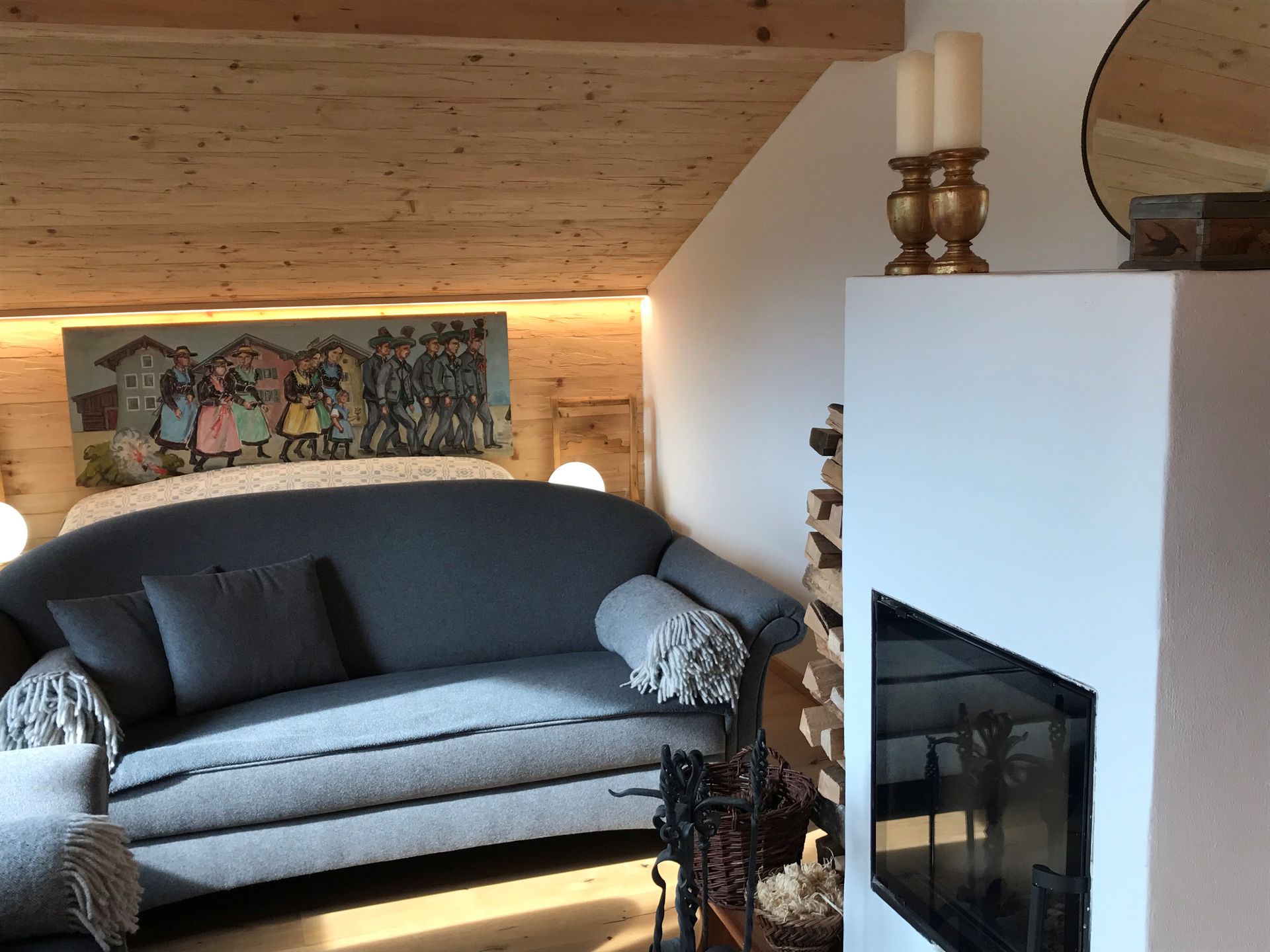 Wohnbereich mit grauem Sofa, Kamin und Gemälde über dem Bett in einem Zimmer mit Holzdecke.