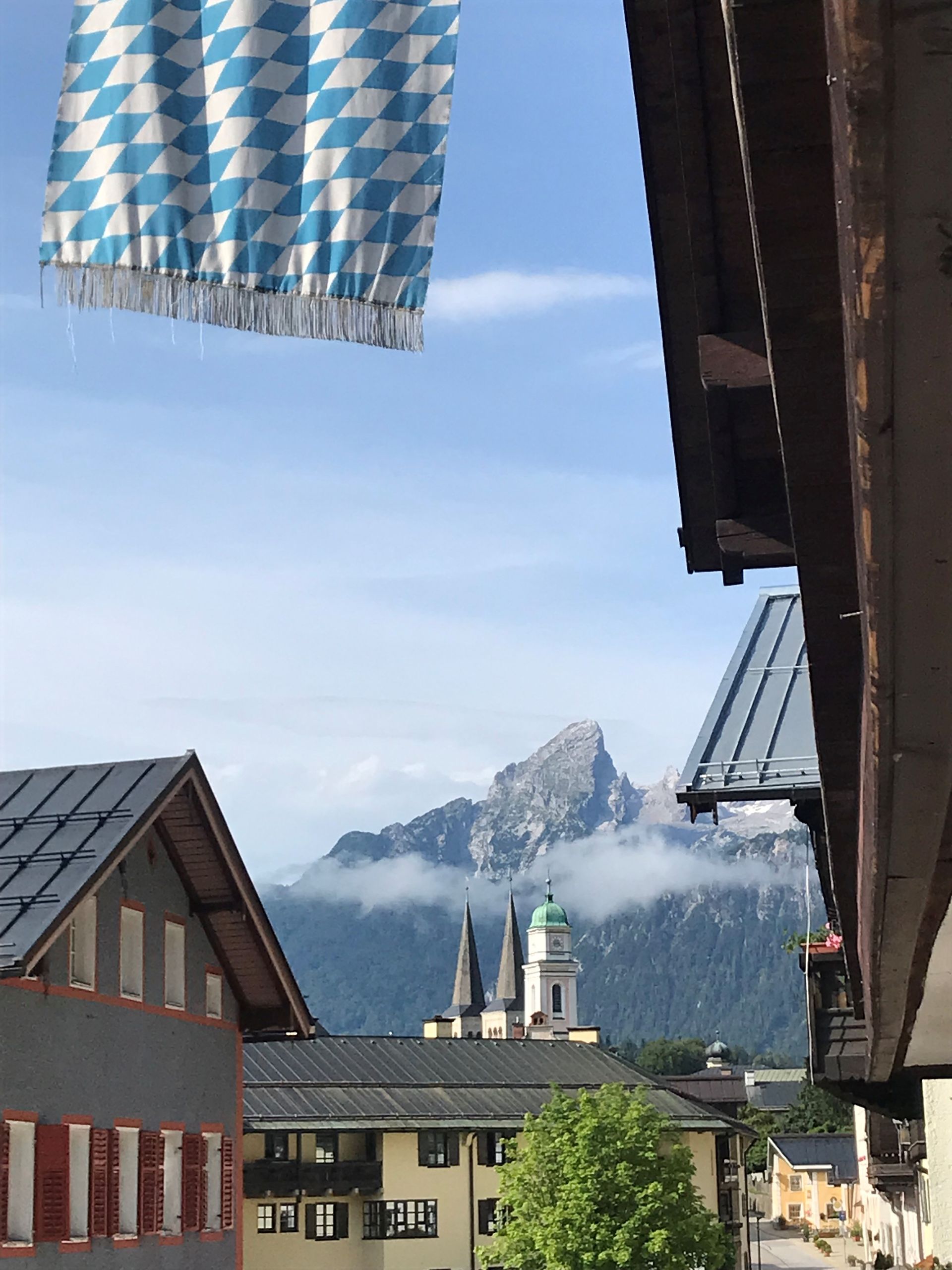 Marktplatz in den bayerischen Alpen. Blau-weiße Flagge, Berggipfel, Kirchtürme, Gebäude.