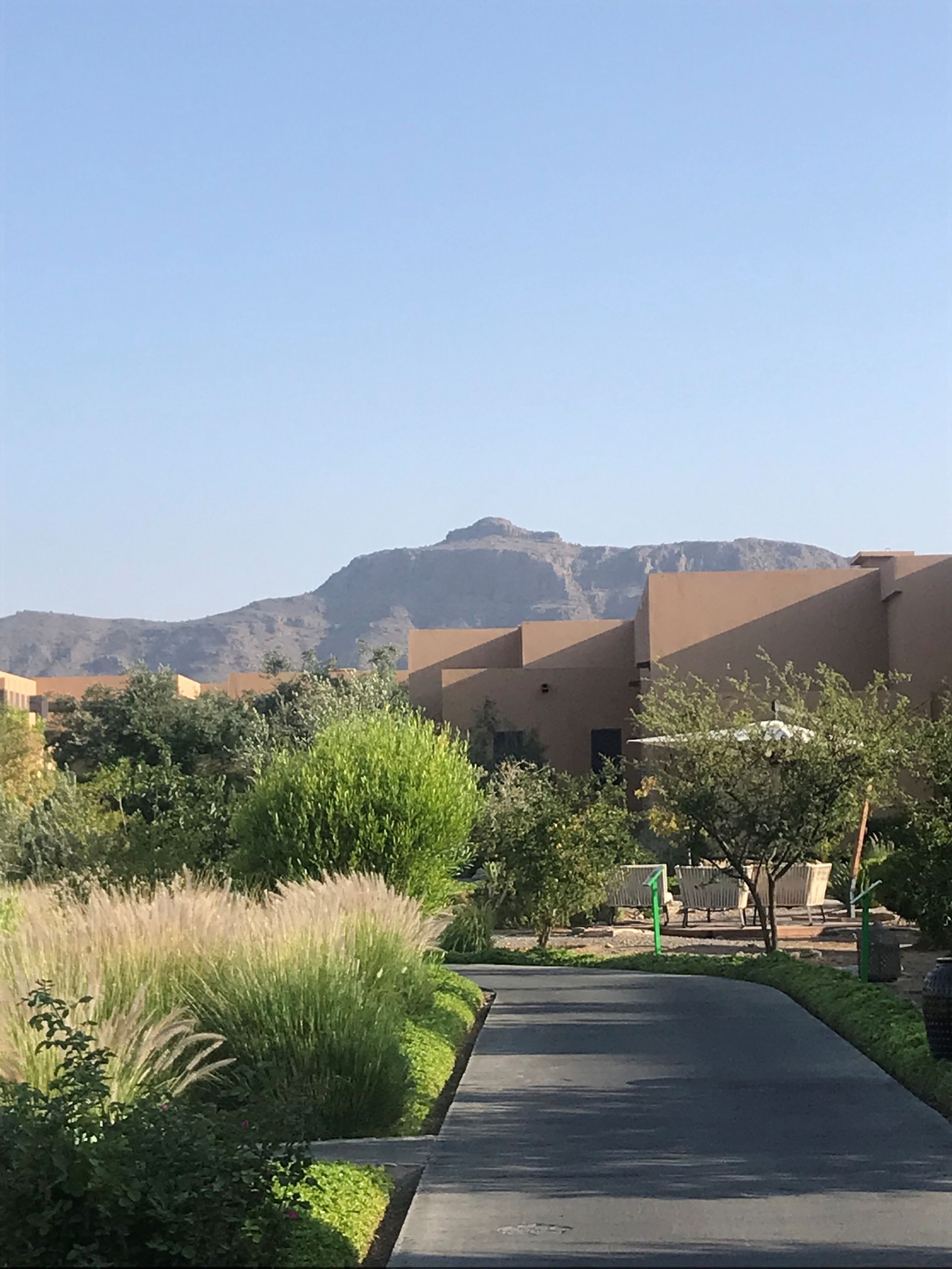 Ein Weg führt durch eine grüne Landschaft, vorbei an beigen Gebäuden und einem fernen Berg unter klarem Himmel.