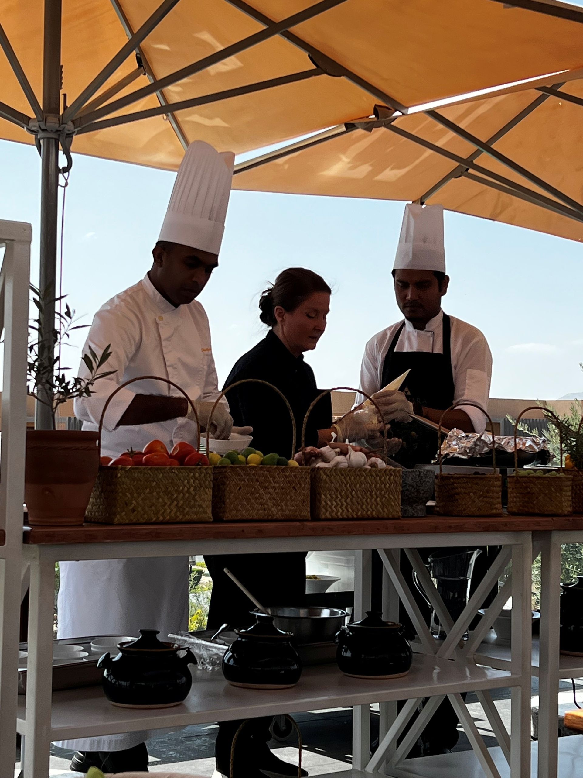 Köche in weißen Uniformen bereiten Speisen an einem Buffet im Freien unter einem Sonnenschirm zu.