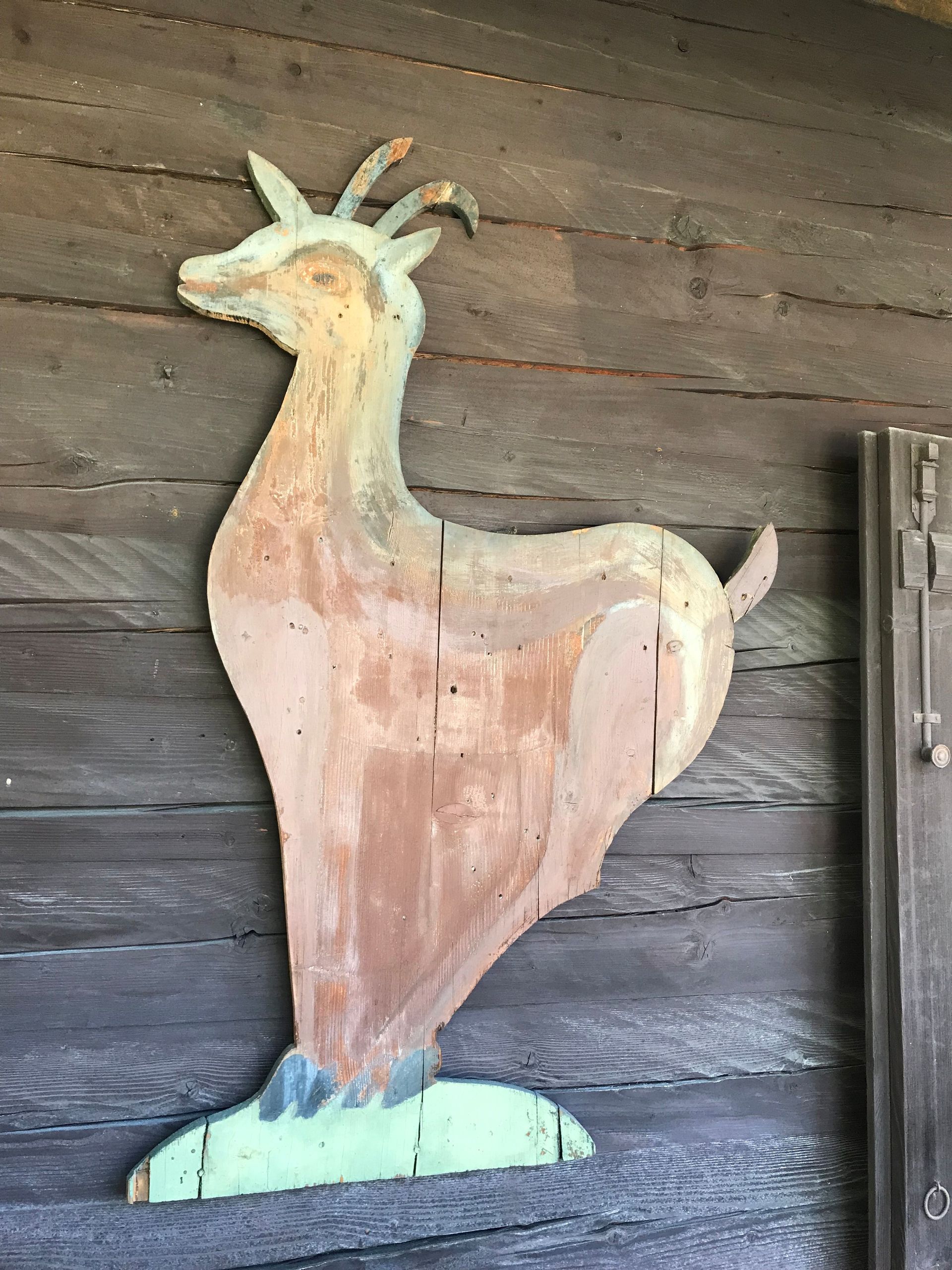 Ausgeschnittene Holzfigur einer Ziege, bemalt in Pastellfarben, angebracht an einer Holzwand.