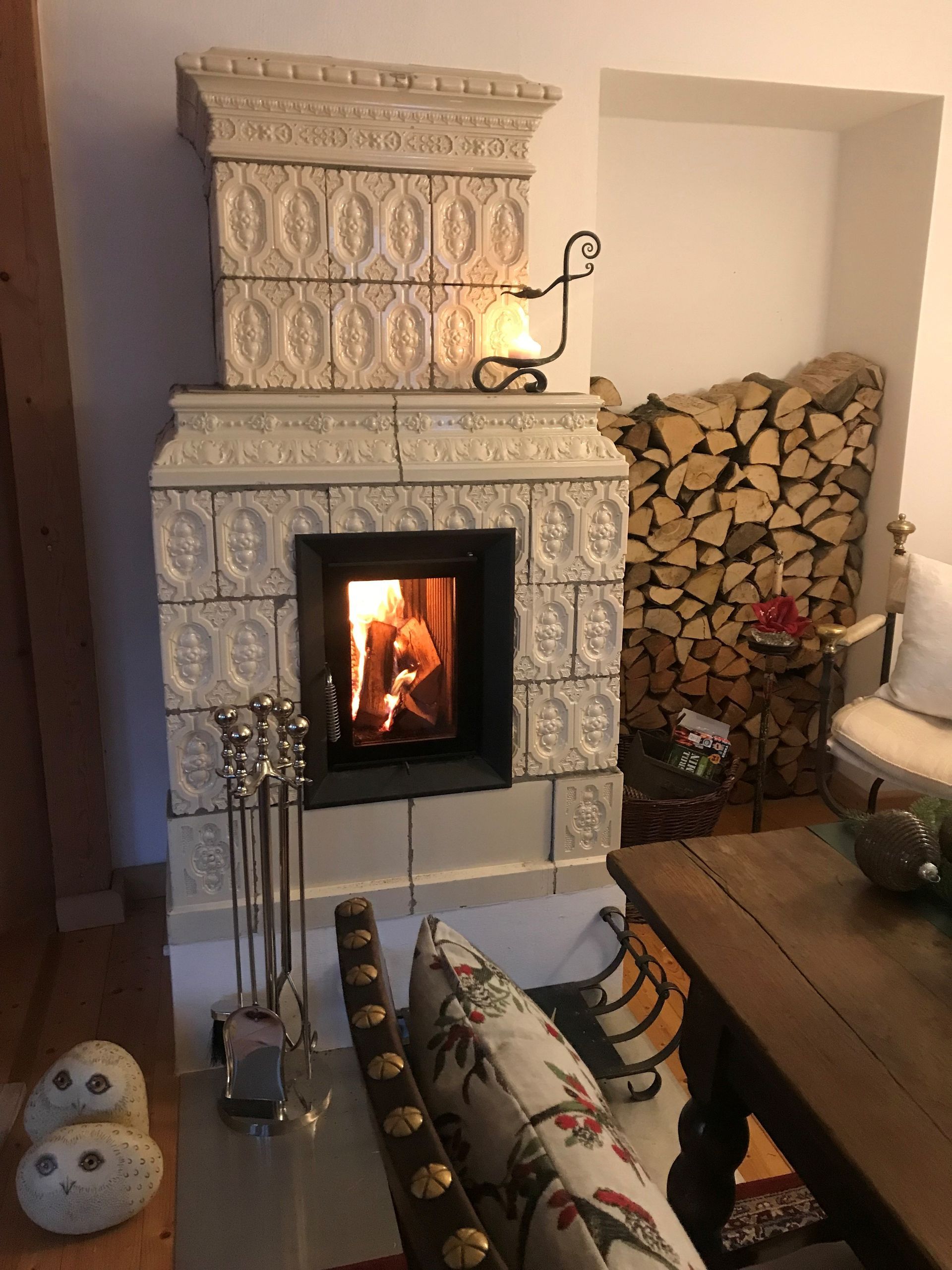 Kamin mit weiß gefliester Umrandung, brennendem Feuer und gestapeltem Brennholz.