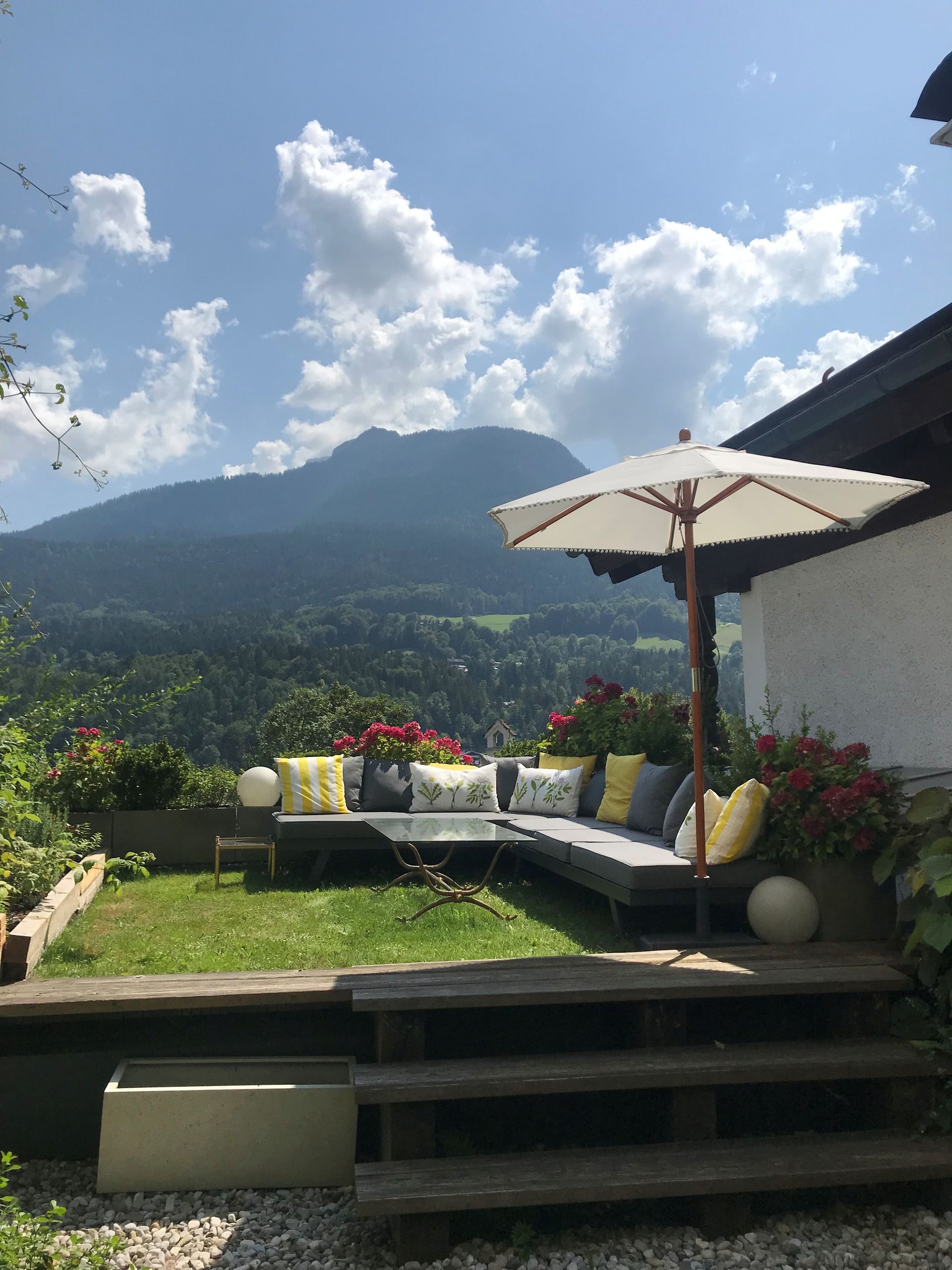 Garten mit Bergblick, Eckcouch, Sonnenschirm, Blumen, grünem Gras und blauem Himmel mit Wolken.
