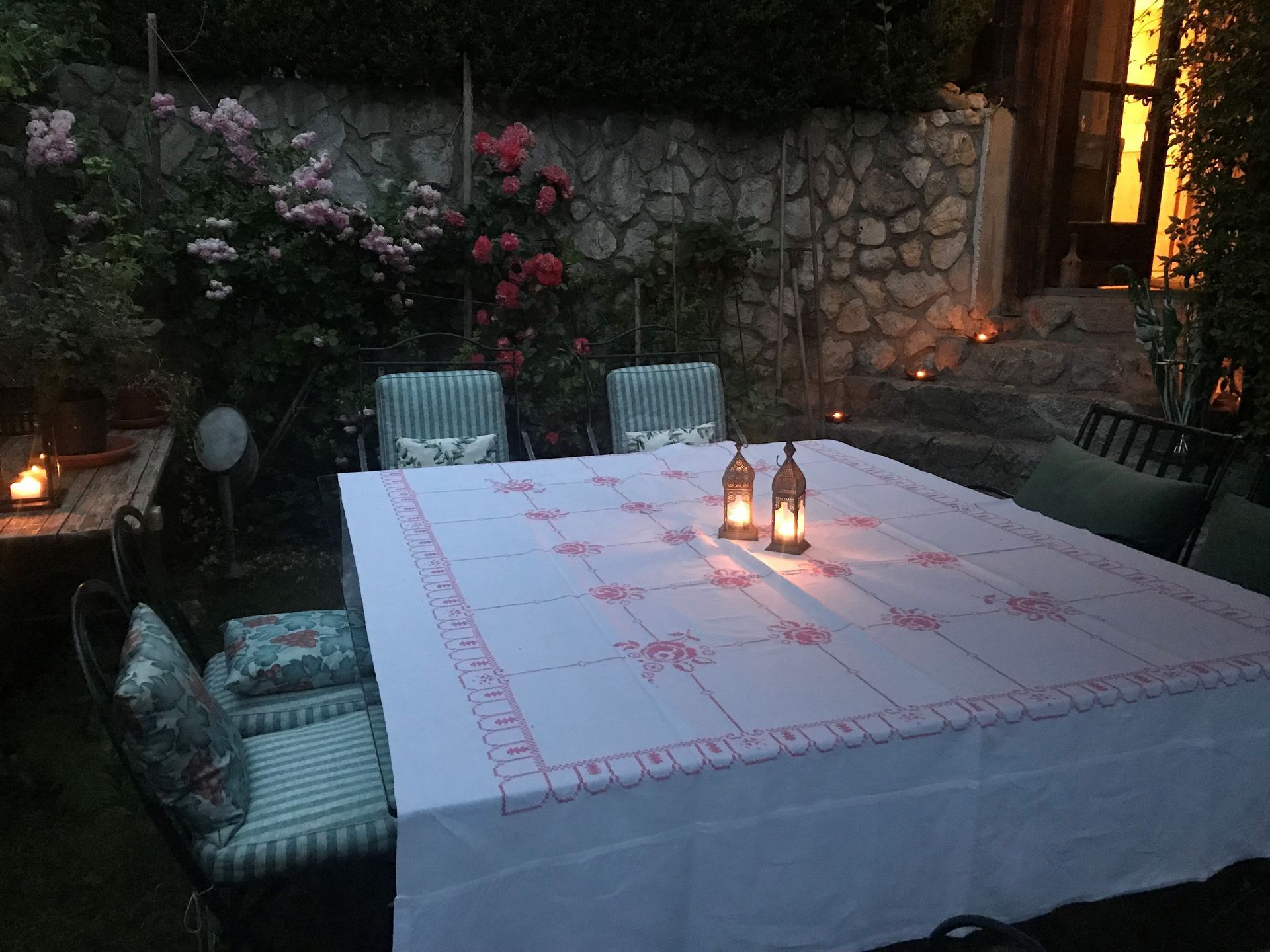 Für den Abend ist ein Esstisch im Freien gedeckt. Brennende Kerzen, Rosen und grüne Stühle.
