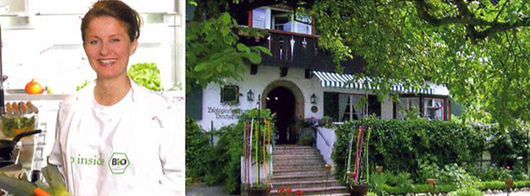 Gabi Kurz im weißen Kittel lächelt neben einem Gebäude, umgeben von Grün.