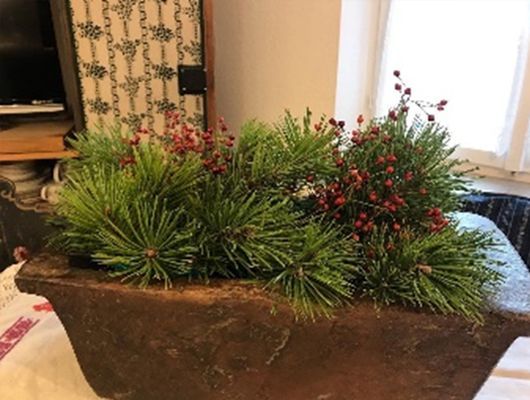 Grüne Kiefer und rote Beeren in einem braunen, rechteckigen Pflanzgefäß auf einem Tisch.