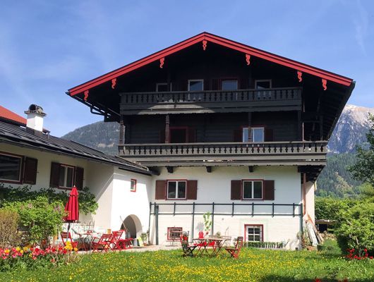 Haus Alpenveilchen - Zweistöckiges Haus mit Holzbalkon und roten Zierleisten vor einer Bergkulisse.