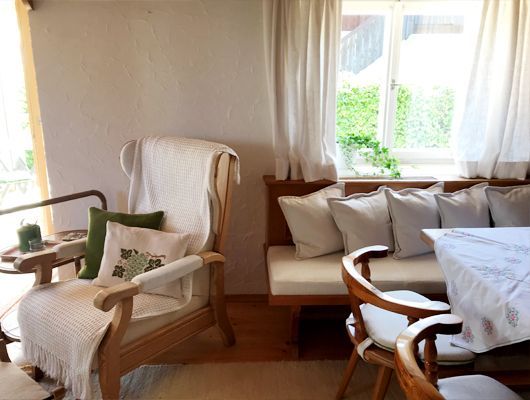 Gemütliches Zimmer mit Holzmöbeln: Stuhl, Bank und Tisch. Weiche weiße Kissen und Vorhänge.