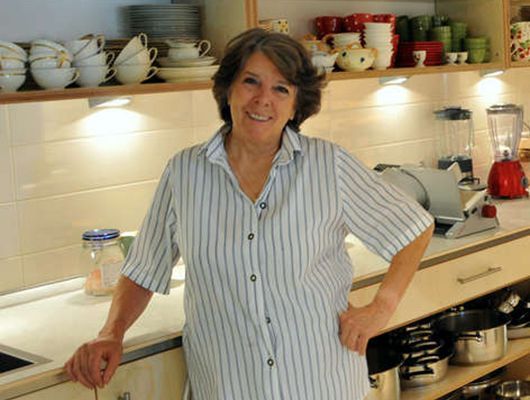 Christl Kurz in einem gestreiften Hemd lächelt in einer Küche mit Geschirr, einem Mixer und Töpfen.