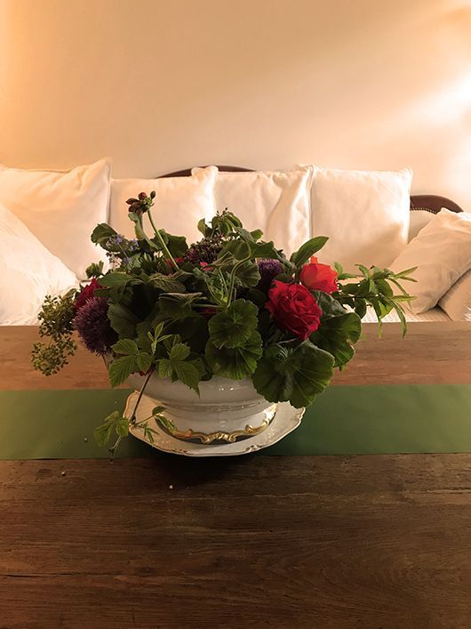 Blumenarrangement in einer weißen Schale auf einem Holztisch, mit weißen Kissen im Hintergrund.
