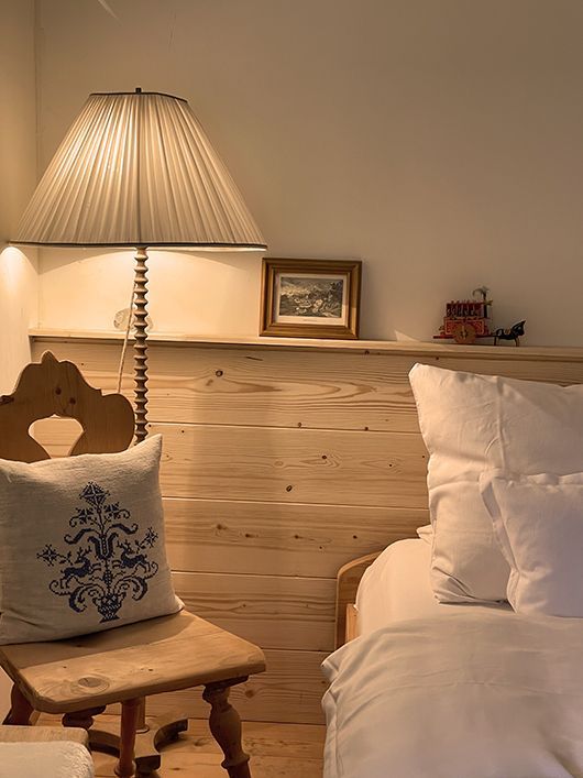 Schlafzimmer mit hellem Holzkopfteil, Stuhl mit dekorativem Kissen, Lampe und weißer Bettwäsche.