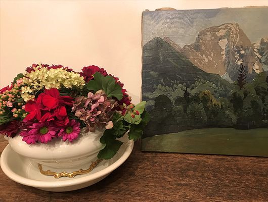Blumenarrangement in einer weißen Schale neben einem Gemälde einer Berglandschaft.