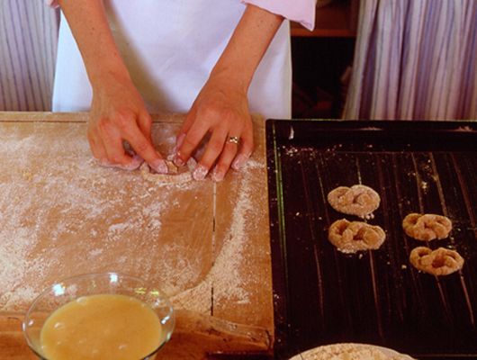 Hände formen Teigbrezeln auf einem bemehlten Holzbrett, neben einem Backblech mit Brezeln.