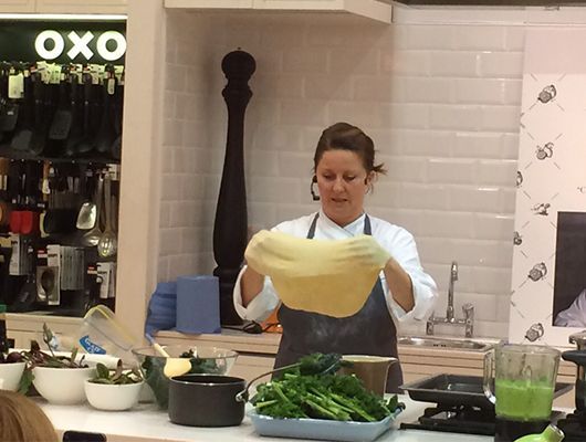 Gabi Kurz in Kochkleidung hält in einer Küche eine große Teigplatte hoch.