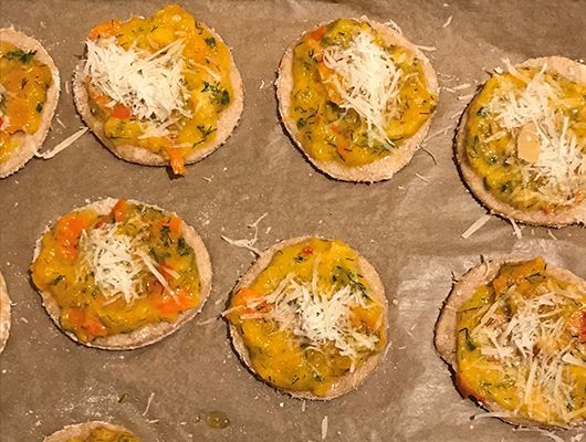 Englische Muffins mit orangefarbenem Gemüsebelag und geriebenem Käse, auf Backpapier gebacken.