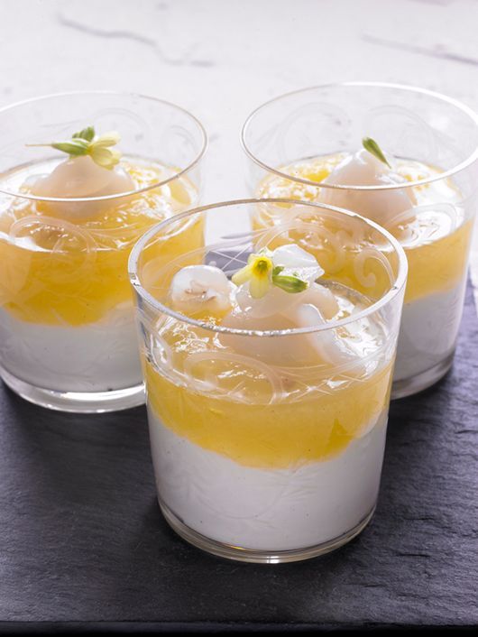 Drei Dessertgläser mit Schichten aus Weiß und Gelb, garniert mit Früchten und Blüten.