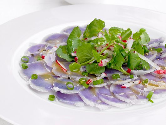 Lila Kartoffelcarpaccio mit Brunnenkresse, Radieschen und Frühlingszwiebeln auf weißem Teller.