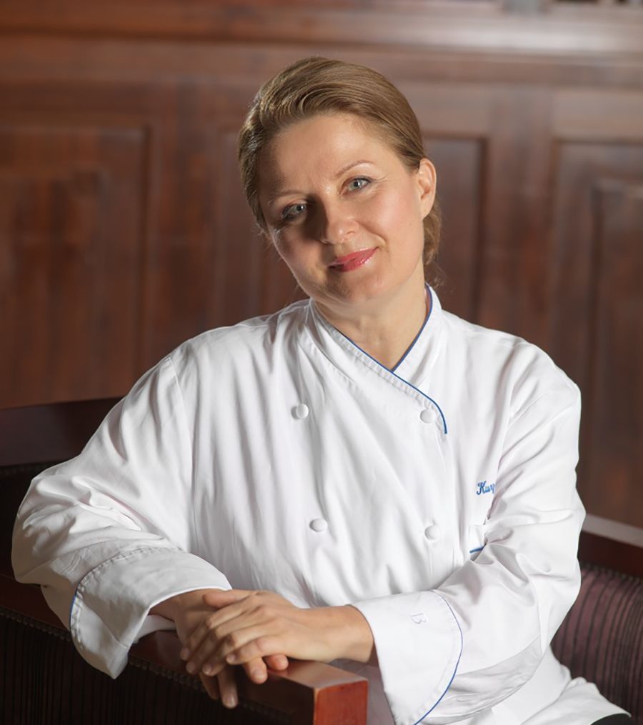 Gabi Kurz in Kochjacke, lächelnd, auf Holzstuhl gelehnt, gegen Holzvertäfelung.