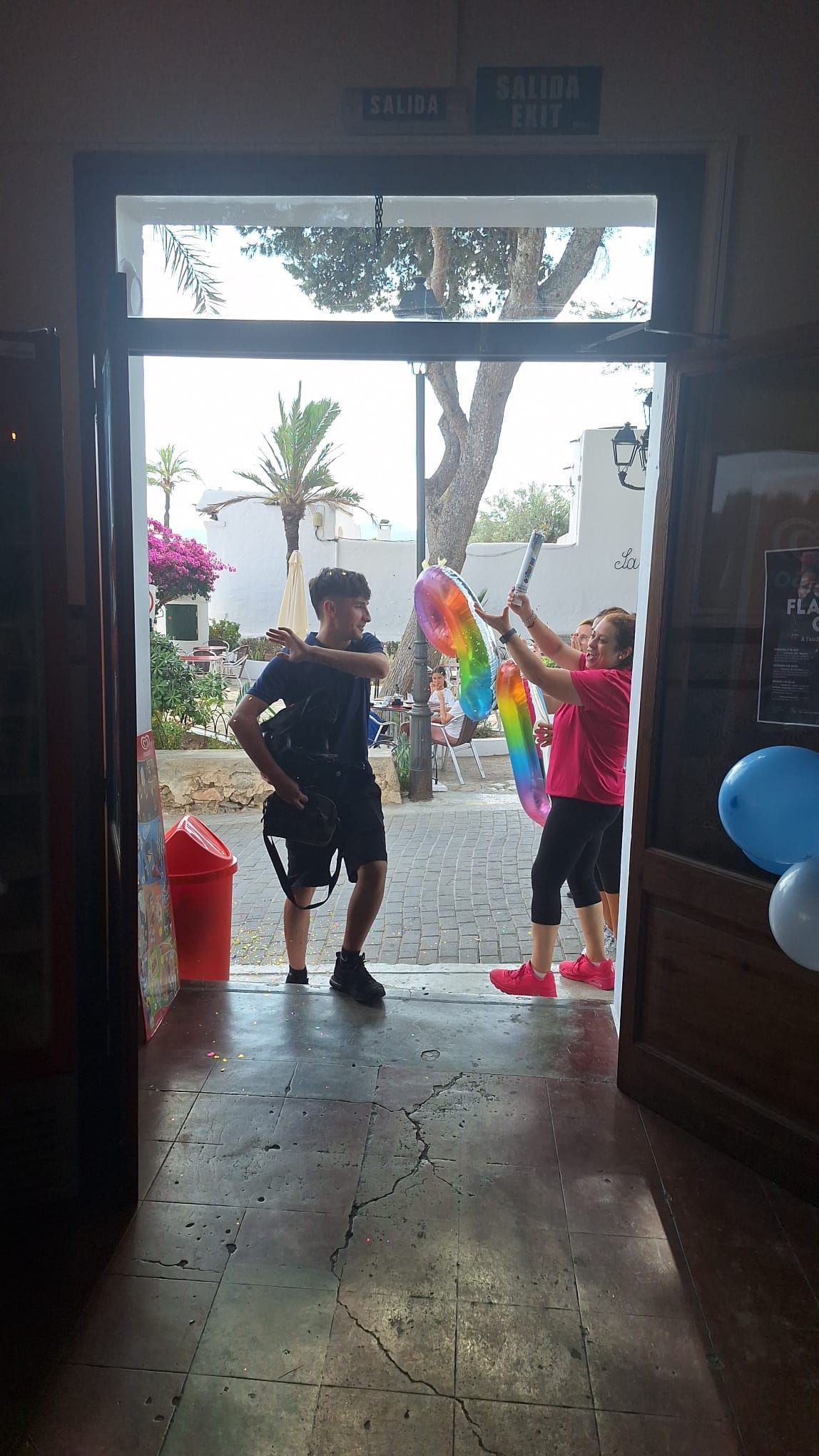 Dos personas están de pie en un portal, una de ellas sosteniendo serpentinas de colores del arcoíris. Afuera, se ve un patio con flores y una palmera.