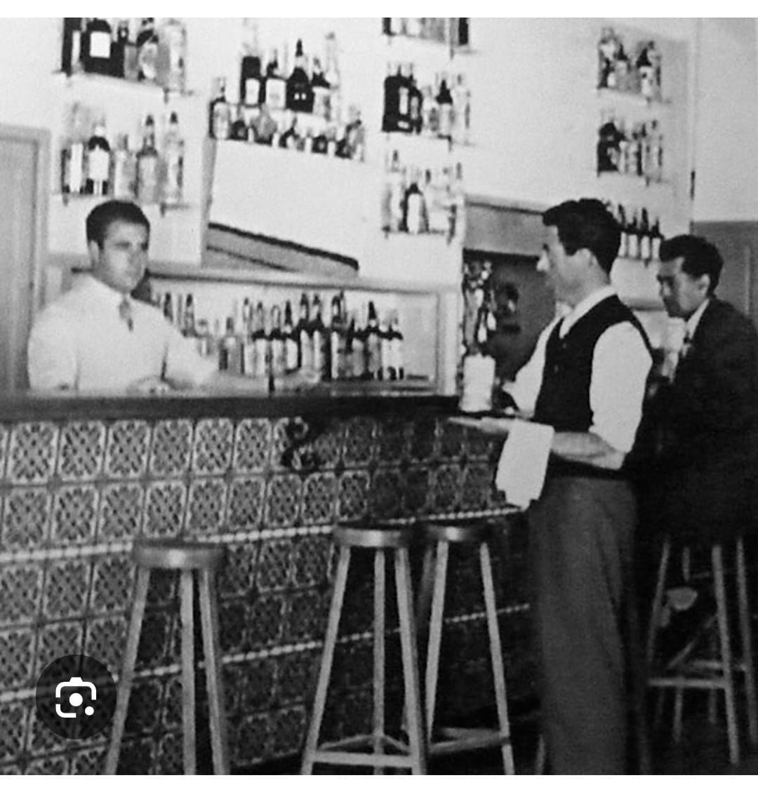 Fotografía en blanco y negro del interior de un bar. Un camarero detrás de la barra, otro atendiendo a un cliente y un cliente en la barra.