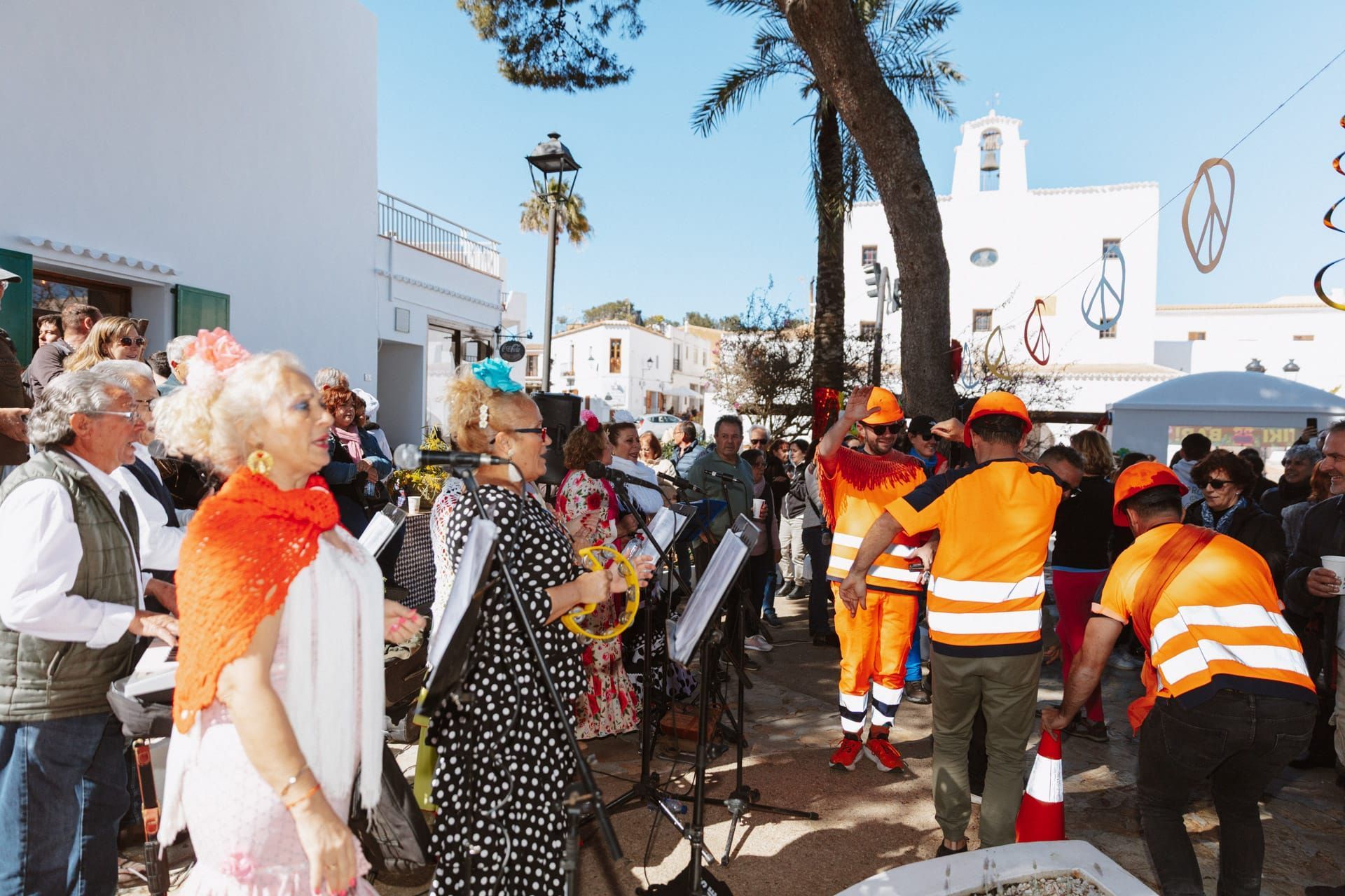 La gente se reunió al aire libre, algunos con instrumentos musicales, cerca de edificios blancos. Había trabajadores con chalecos naranjas y uniformes de construcción.