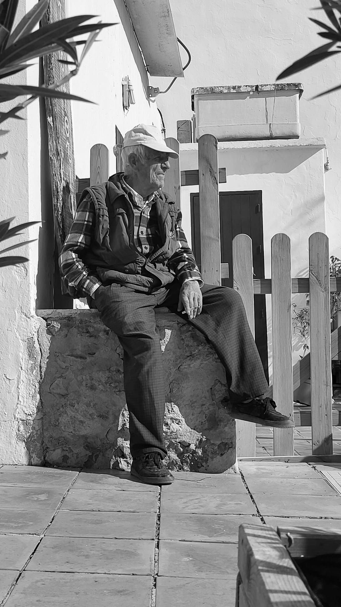 Un hombre mayor, con gorra, sentado en una repisa de piedra, apoyado contra una valla de madera.