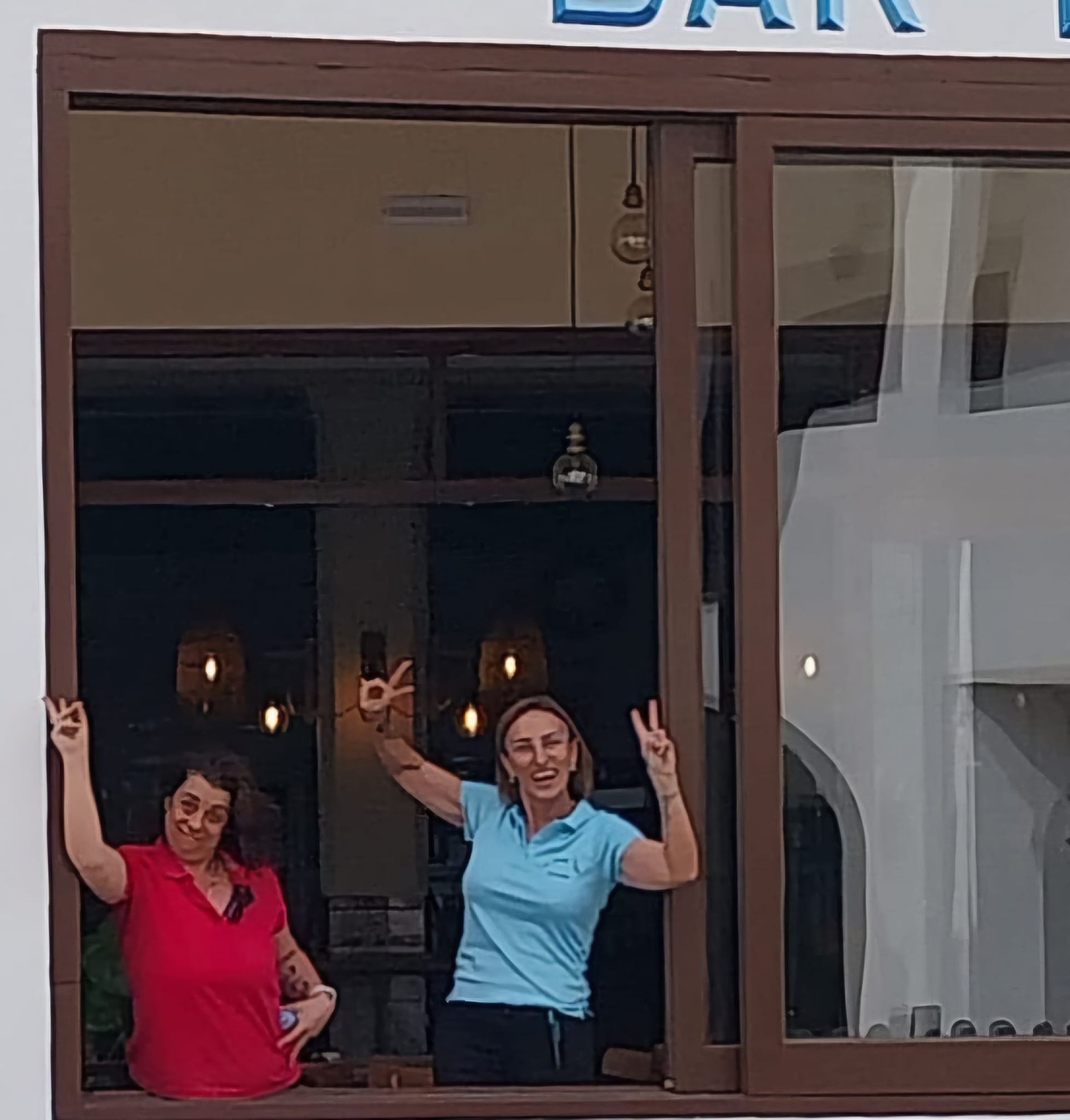 Dos mujeres en un escaparate haciendo la señal de la paz. Una viste de rojo, la otra de azul, ambas sonriendo. Escenario exterior, posiblemente un bar.