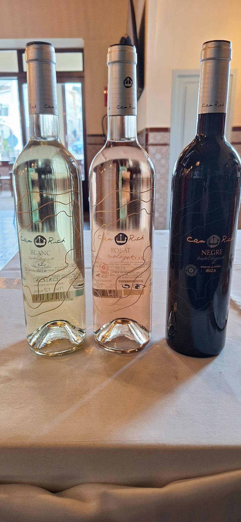 Tres botellas de vino, una blanca, otra rosa y otra roja, sobre una mesa. Tapas plateadas y bases decorativas.