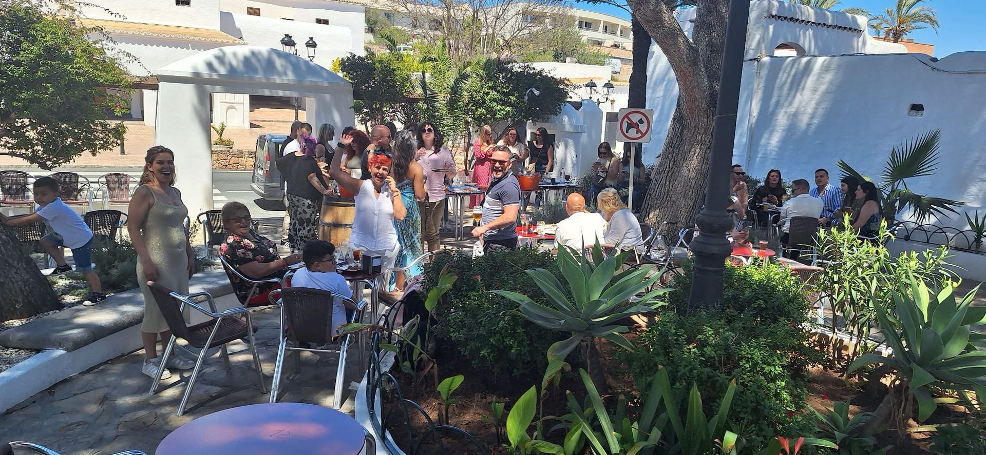 Un grupo de personas se reunió al aire libre en un café. Algunos estaban sentados en mesas, otros de pie, algunos vestidos elegantemente. Edificios blancos al fondo.