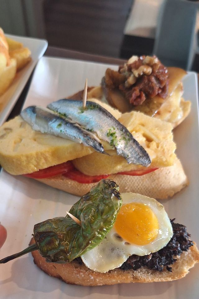 Varias tapas en un plato blanco: una rodaja de sardina y tomate sobre pan; un huevo frito y pimiento sobre pan; y otros aperitivos variados.