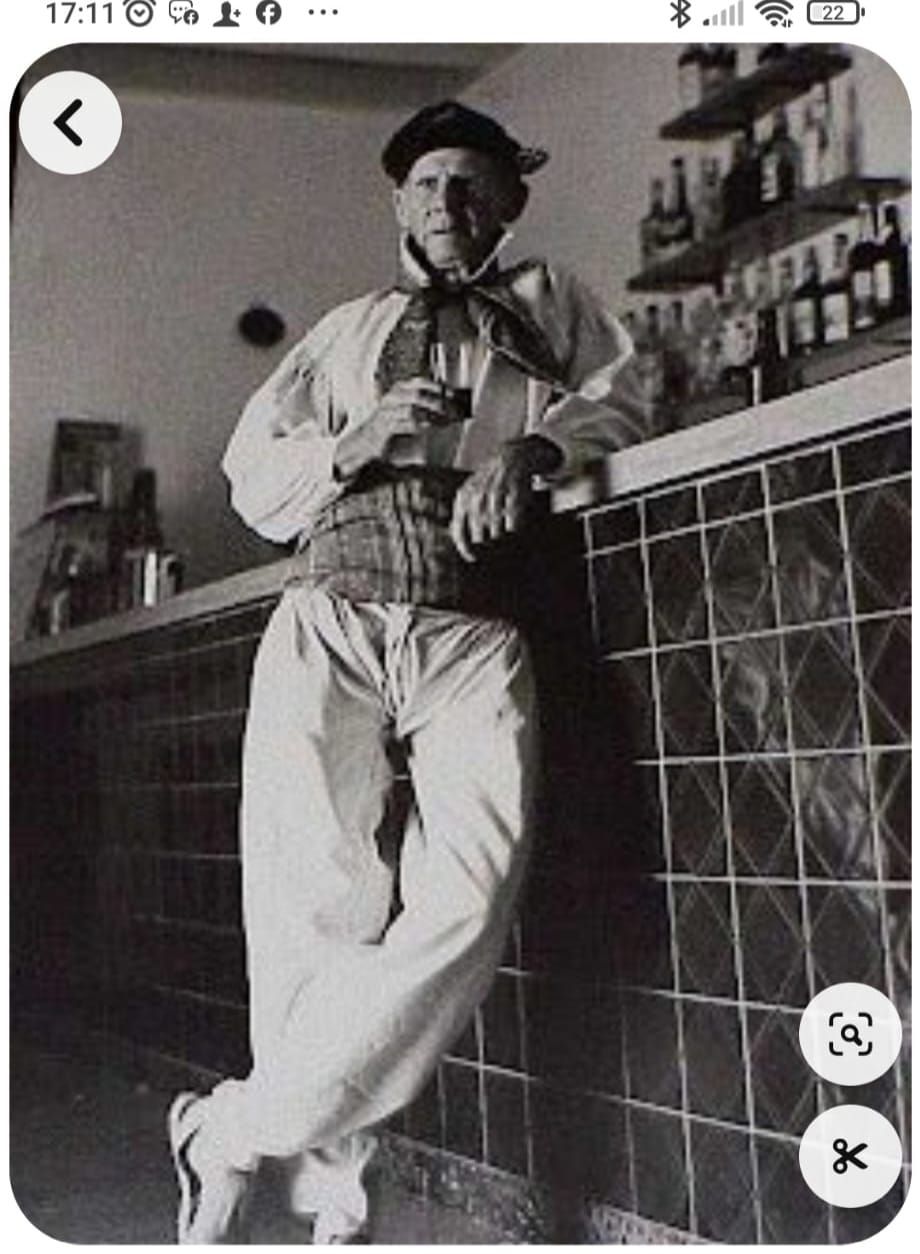 Un hombre con traje tradicional se apoya en una barra. Sostiene una bebida, posando frente a estanterías llenas de botellas.
