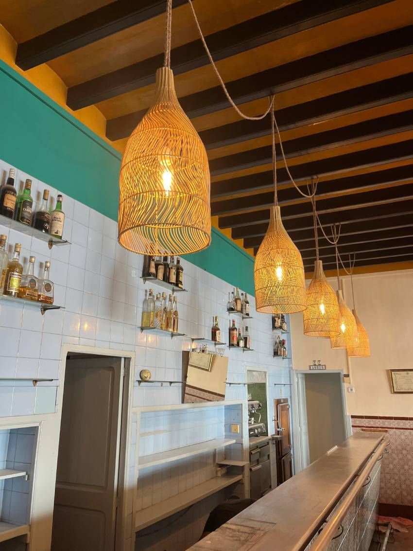 Interior de bar con luces colgantes tejidas sobre un mostrador, botellas exhibidas en estantes contra una pared de azulejos blancos.