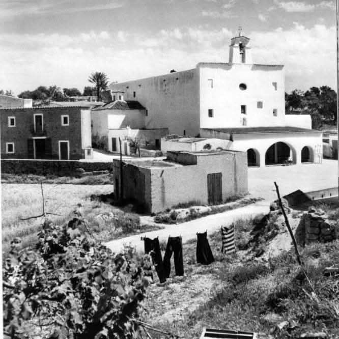 Fotografía en blanco y negro de un edificio blanco con un campanario, posiblemente una iglesia, junto a edificios más pequeños y ropa secándose en un entorno rural.