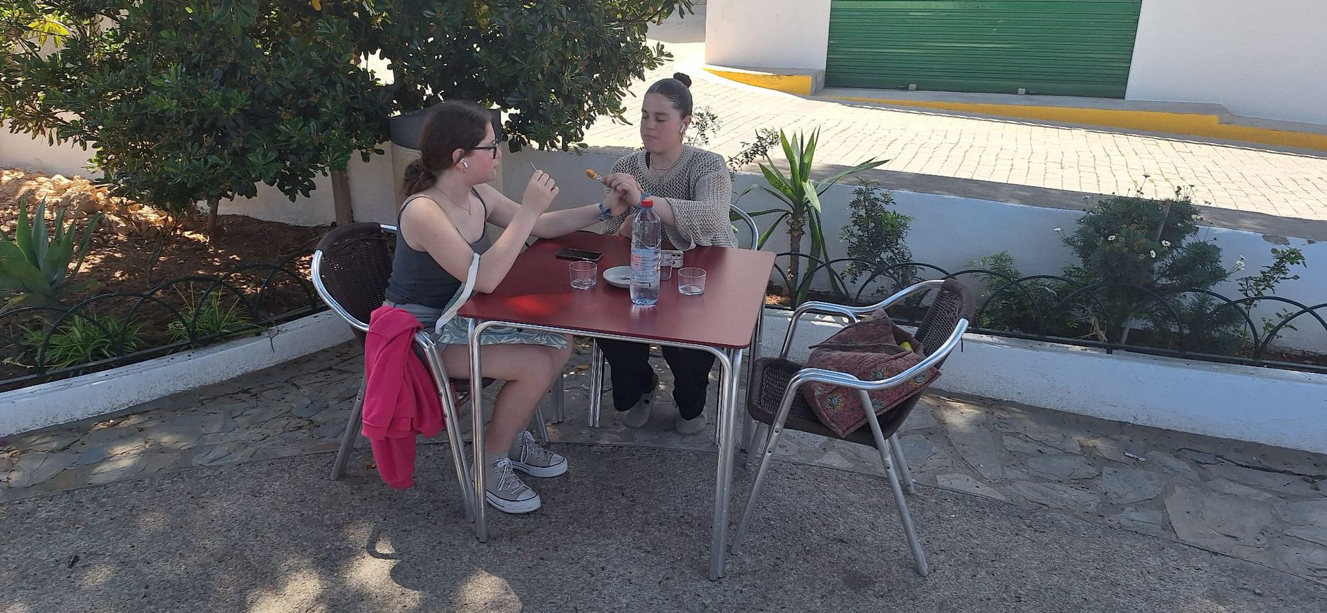 Dos mujeres conversan sentadas en una mesa al aire libre. Una lleva una blusa y falda grises, y un suéter rosa en la silla. La otra lleva una camisa estampada.