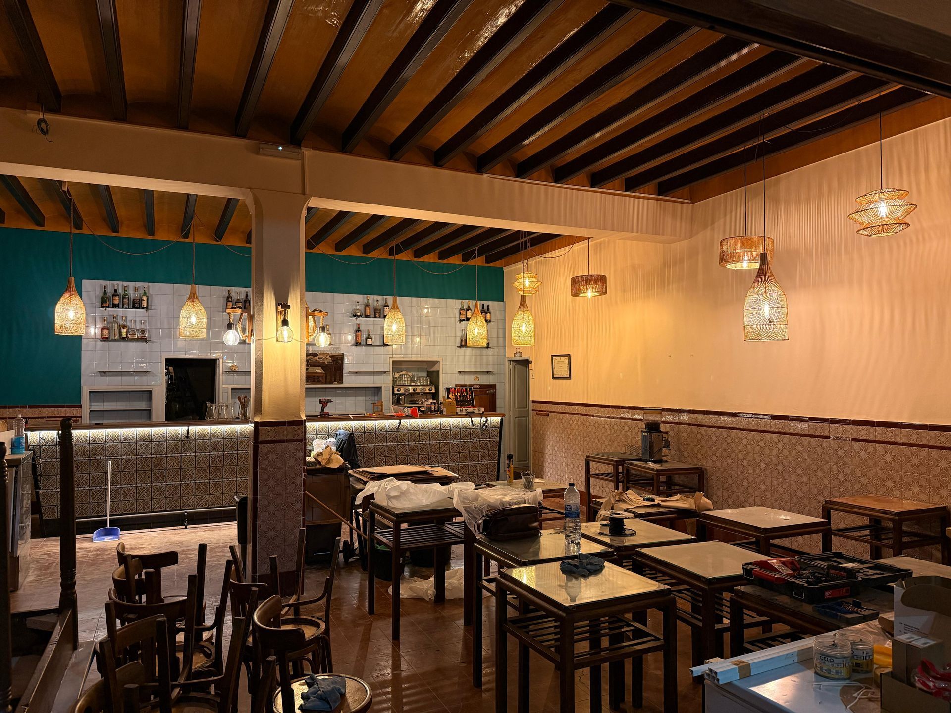 Interior de un restaurante con mesas, sillas y barra. Iluminación cálida con paredes en tonos verde azulado y beige.