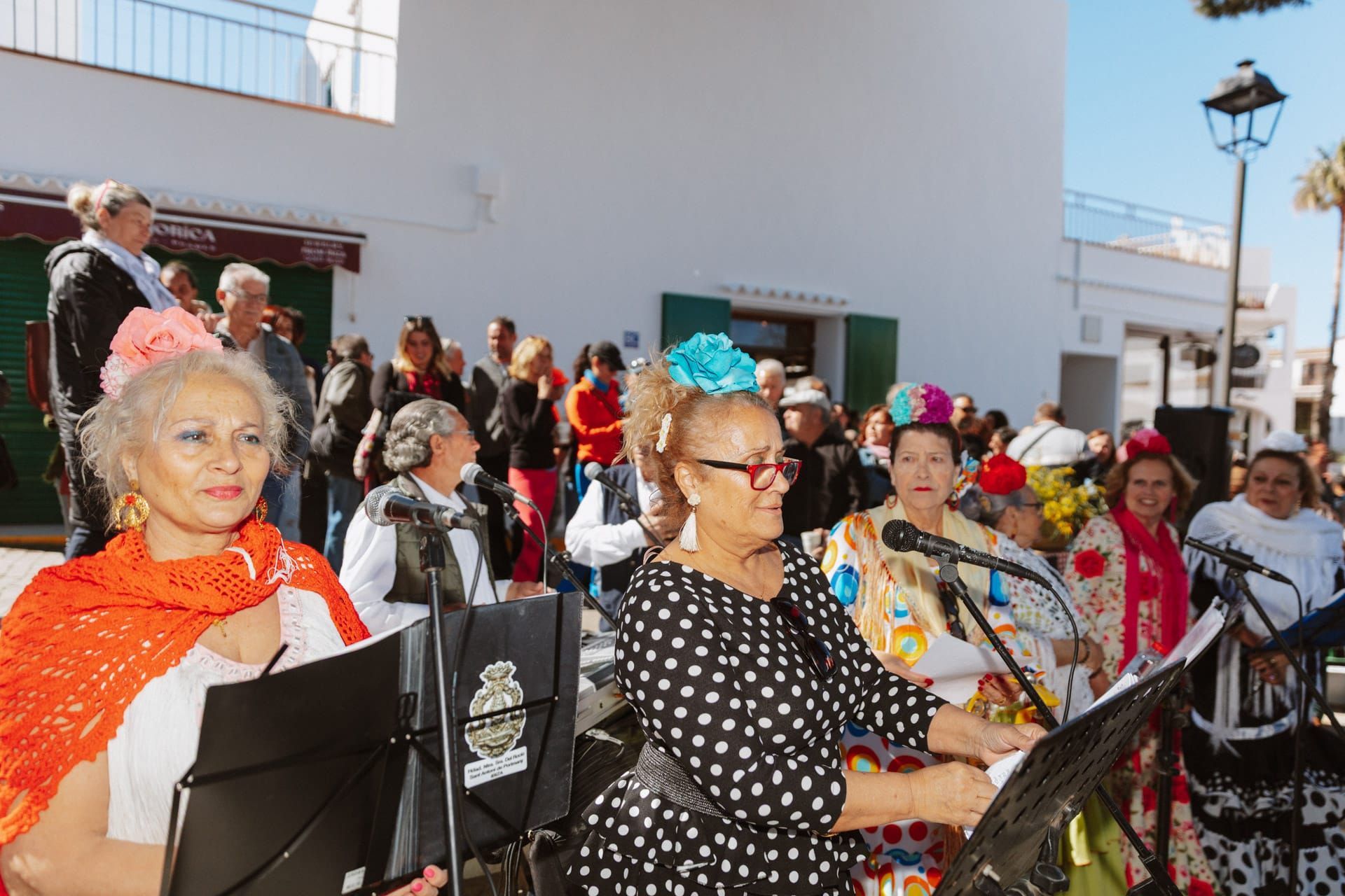 Un grupo de mujeres con atuendos coloridos canta en un evento al aire libre. Algunas llevan tocados florales y sostienen partituras.