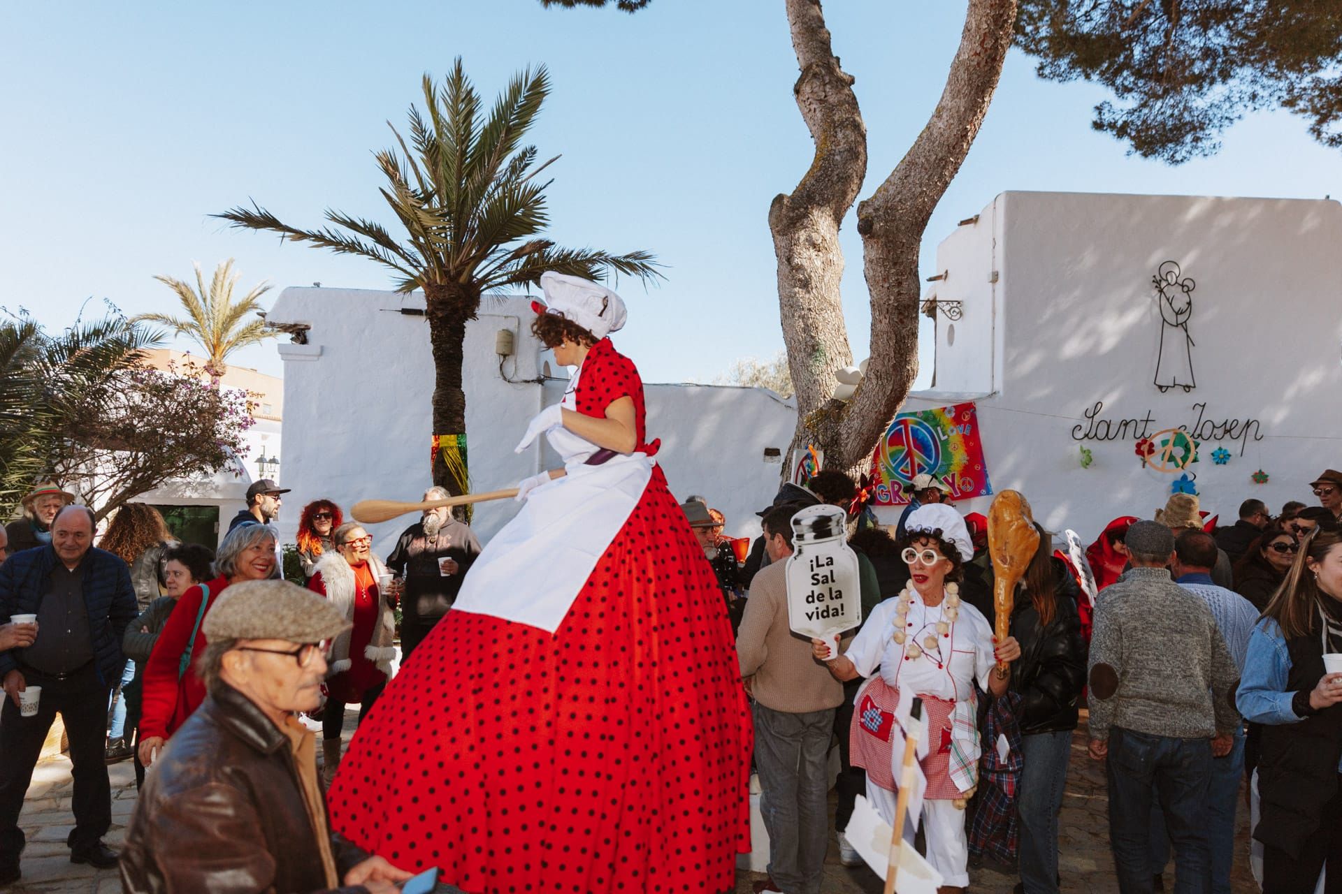 Un zancudo con un vestido rojo y un delantal blanco actúa en un festival, con una multitud de personas disfrazadas al aire libre.