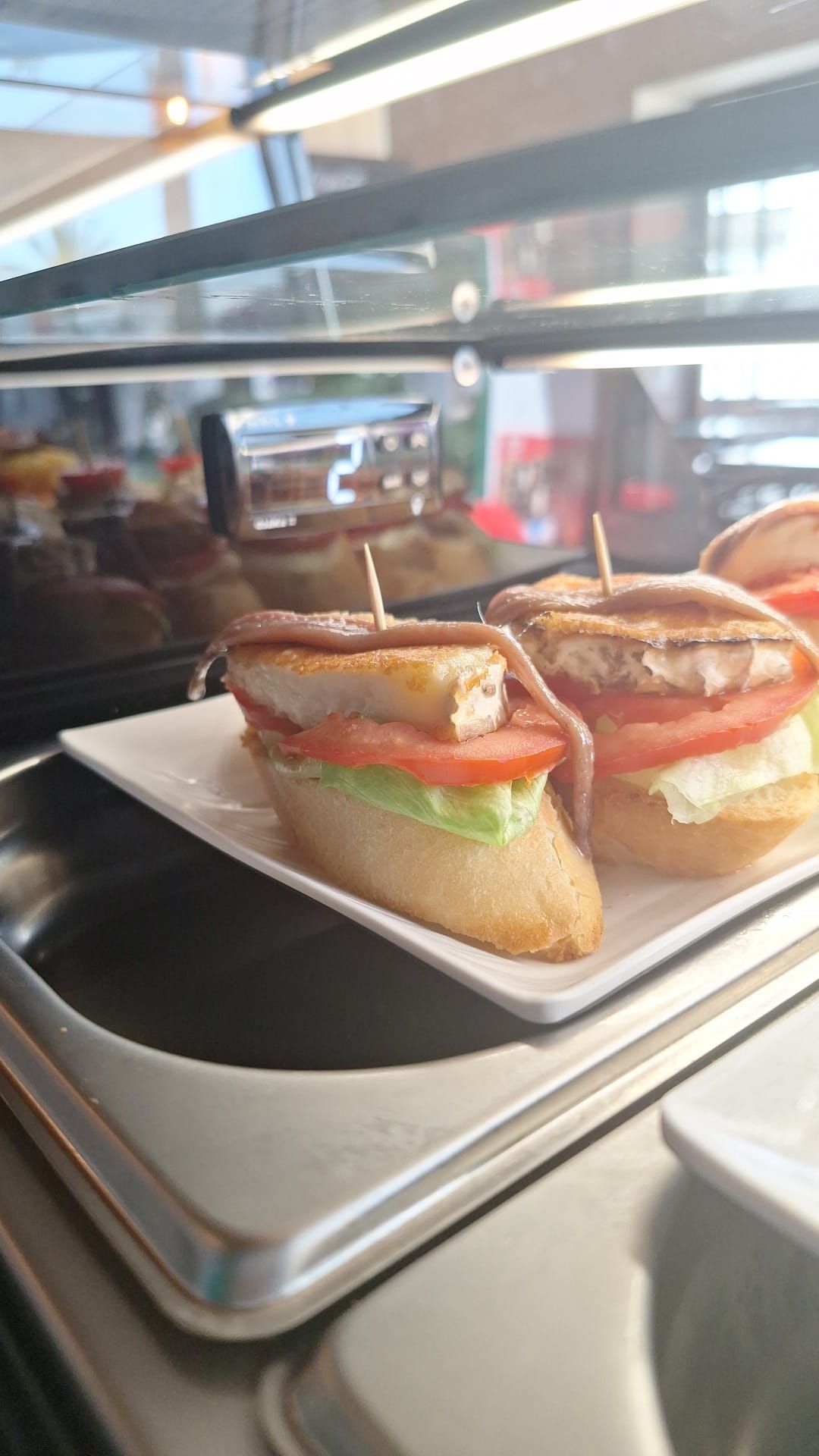 Sándwiches pequeños en un plato blanco, en una vitrina. Cada sándwich lleva una rodaja de tomate y un trozo de carne.