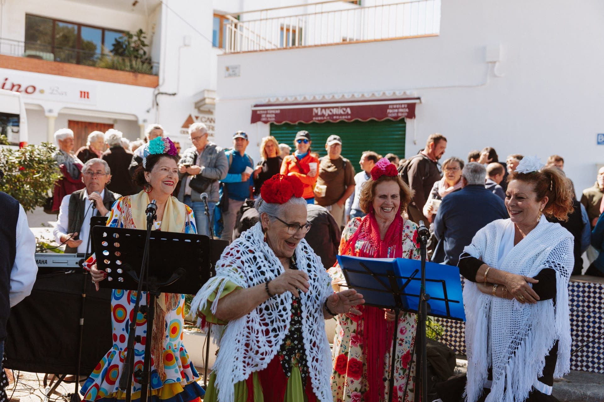 Un grupo de mujeres con coloridos trajes de flamenca canta y actúa al aire libre. Expresiones alegres; público visible al fondo.