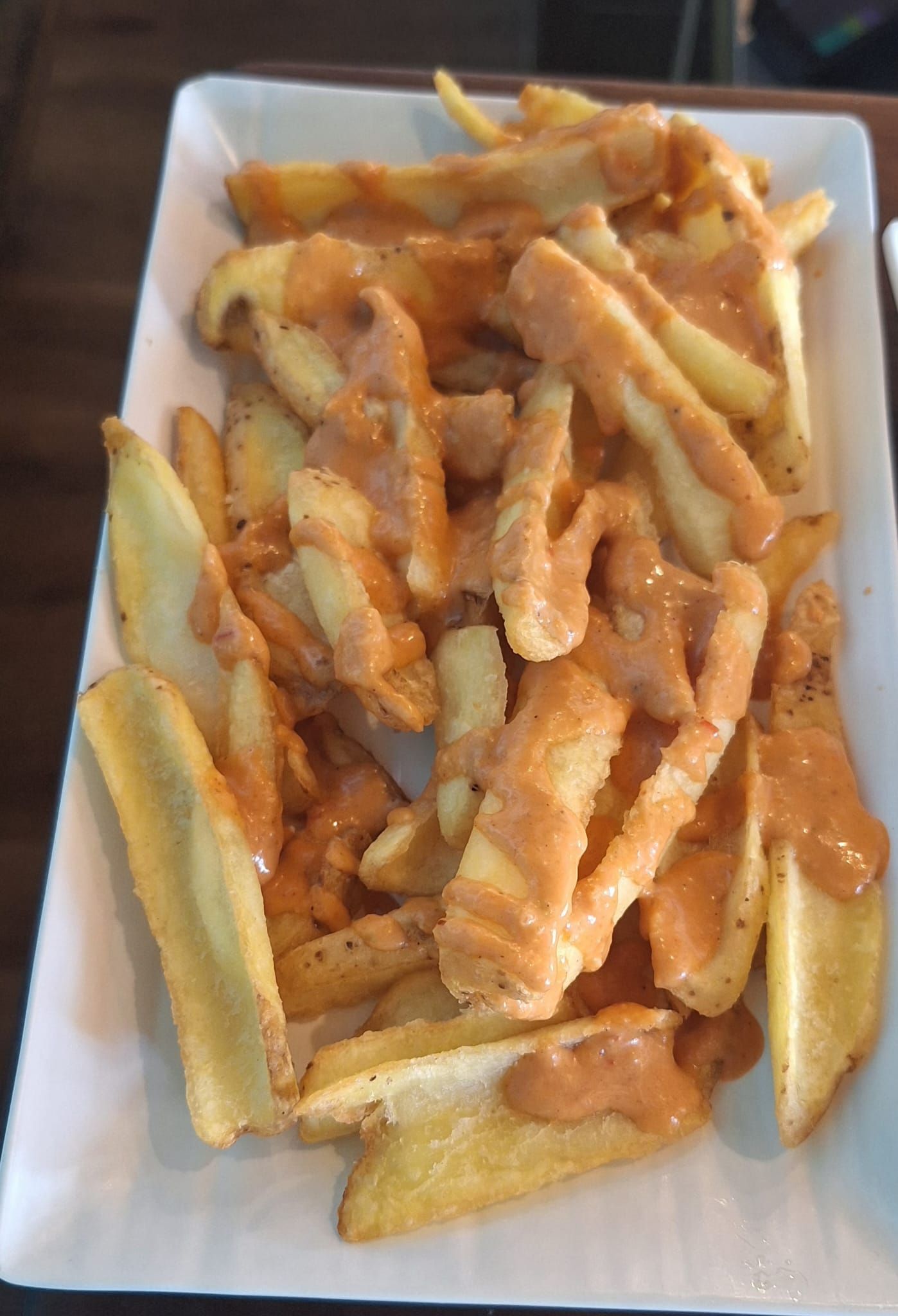 Plato de papas fritas doradas rociadas con una salsa cremosa de color marrón rojizo, probablemente alioli o un condimento similar, servidas en un plato rectangular blanco.