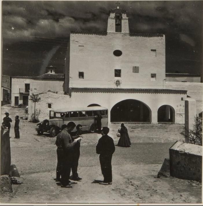 Fotografía en blanco y negro de una iglesia de estuco blanco con una entrada arqueada, gente caminando enfrente y un autobús estacionado cerca.