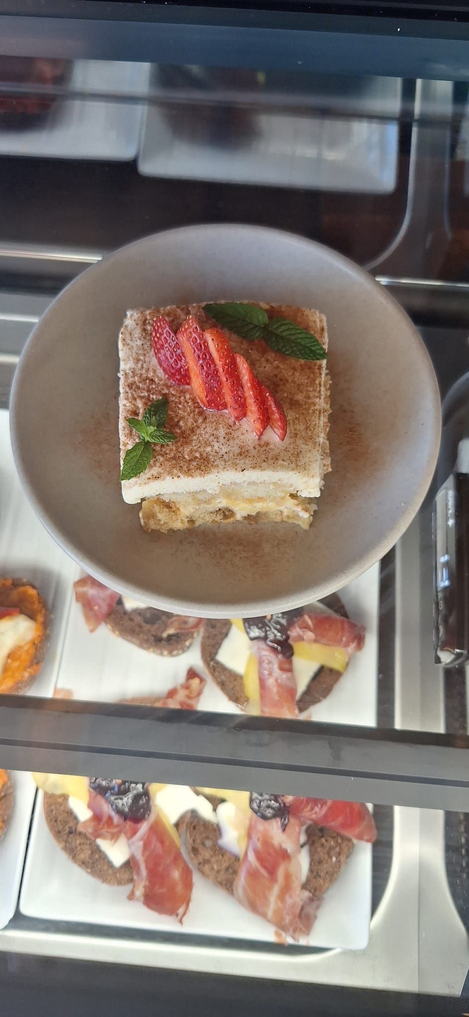 Un postre cuadrado en un plato, cubierto con fresas rebanadas y menta. Abajo, tapas con carne y queso.