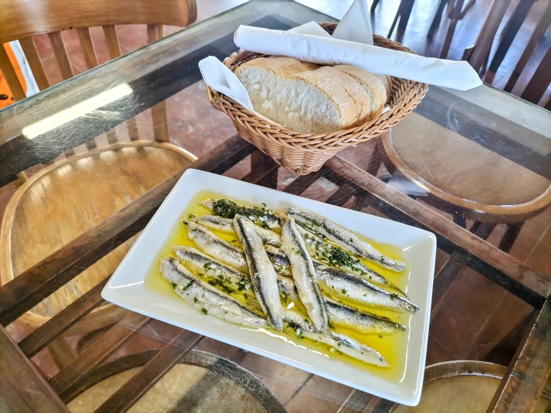 Anchoas en aceite en un plato blanco, con una cesta de pan sobre una mesa.