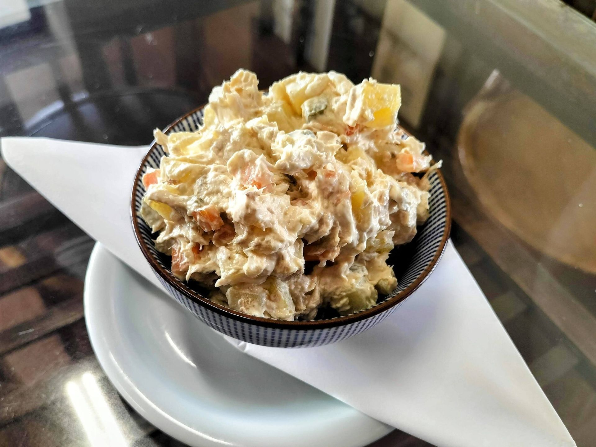 Ensalada de patata en un bol estampado, sobre una servilleta y un plato blanco pequeño. La ensalada es cremosa y está repleta de trozos de patata.