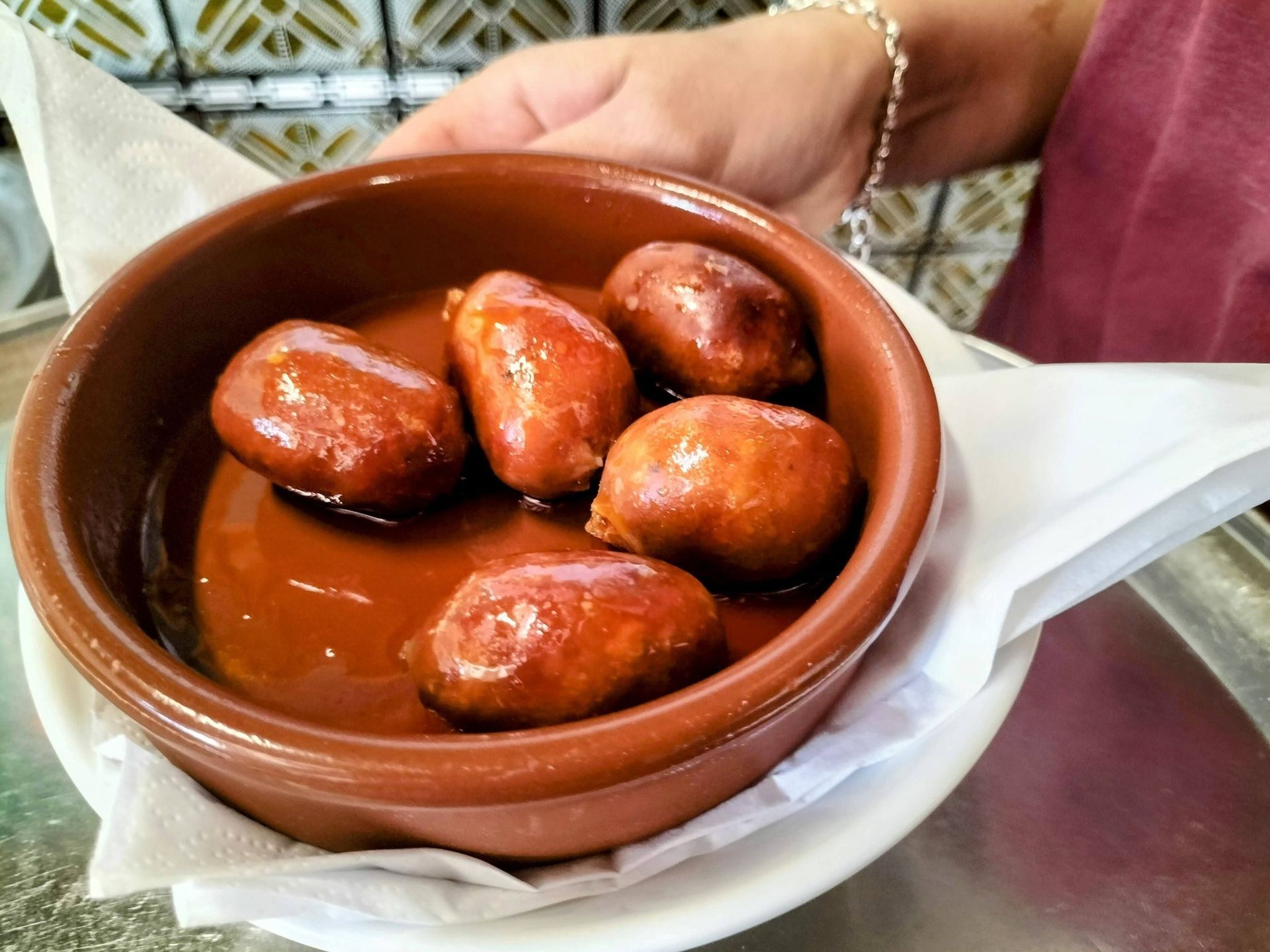 Una mano sostiene un pequeño tazón marrón de chorizos fritos. Los chorizos son de un marrón brillante y reposan en un charco de aceite.