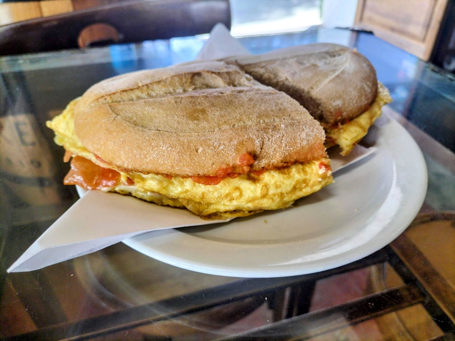 Sándwich de tortilla cortado por la mitad en un plato blanco; el pan es de color marrón claro, el relleno es amarillo y se ve un poco de salsa roja.