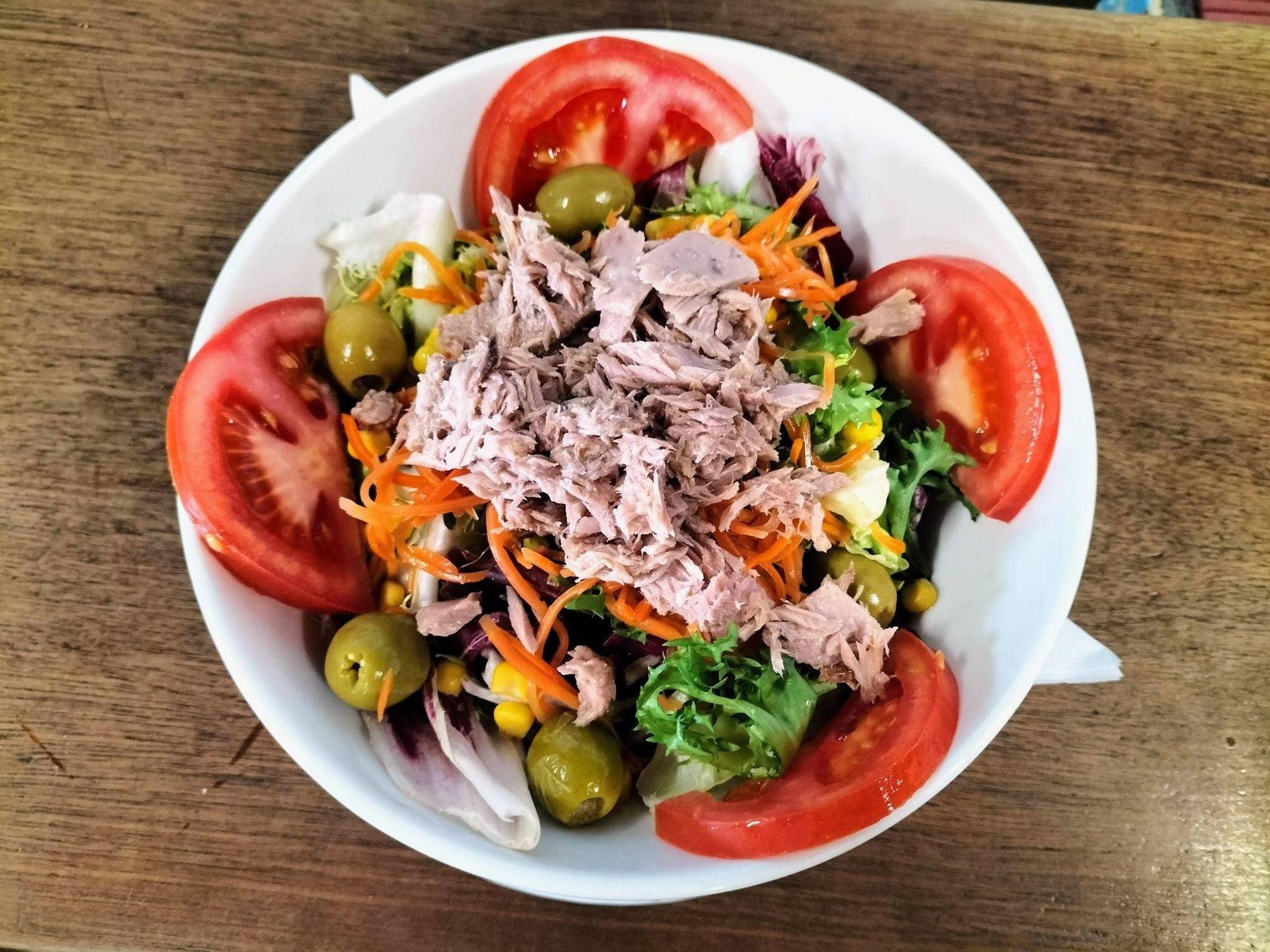 Ensalada de atún con rodajas de tomate y aceitunas en un recipiente blanco sobre una mesa de madera.