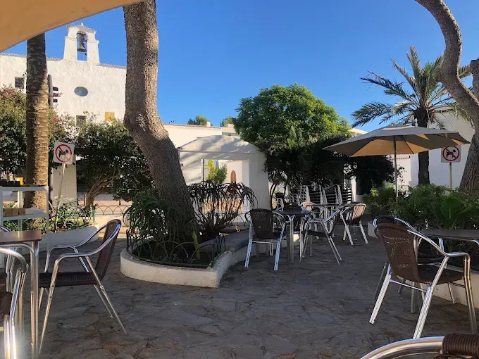 Café al aire libre con mesas y sillas, a la sombra de árboles y sombrillas, frente a un edificio blanco con un campanario bajo un cielo azul.