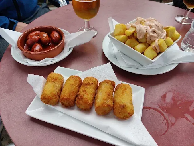 Mesa de tapas españolas: patatas con salsa, chorizo y croquetas. Sobre la mesa se ven una cerveza y un vaso de agua.