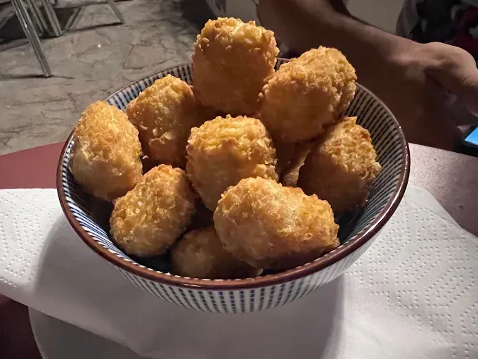 Un tazón de croquetas de patata fritas y doradas reposa sobre una servilleta blanca. Al fondo se ven el brazo y la mano de alguien.